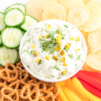 dill pickle dip with pretzels, peppers, chips and cucumbers surrounding it